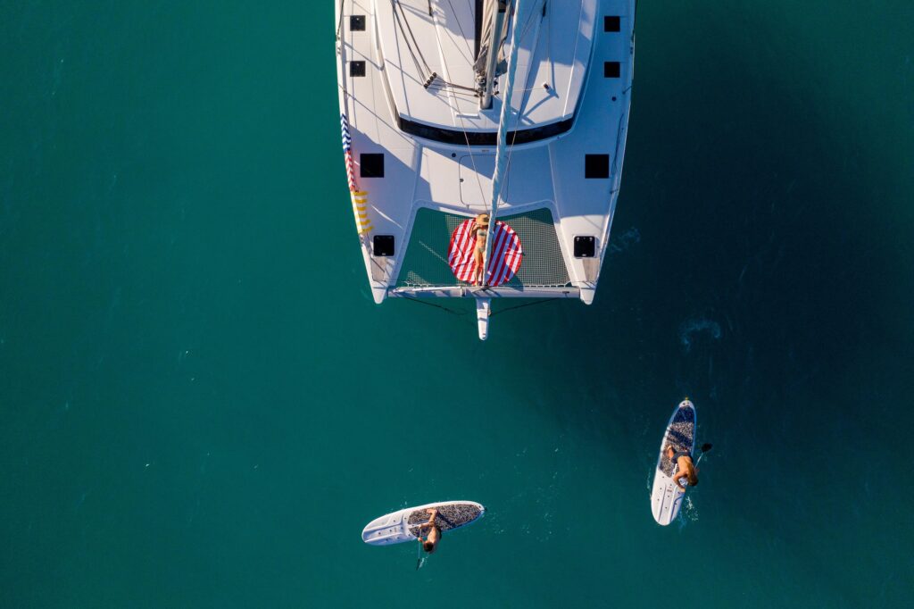 Aerial view of a woman sunbathing on a catamaran in The Whitsundays, with paddle boarders nearby on the clear waters.