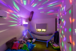 A sensory room with dim lighting, colourful projected lights creating patterns on the walls and ceiling. The room features a hammock, soft cushions, toys, and a calming atmosphere designed for relaxation and stimulation. A small window lets in some natural light, adding to the tranquil setting.