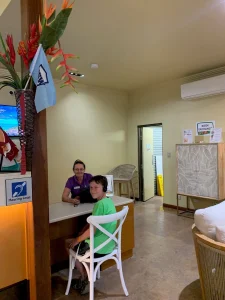 A young boy sitting at a small desk, smiling at the camera, while a staff member in a purple uniform sits across from him with a welcoming expression.