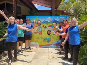 A group of smiling individuals posing enthusiastically around a colourful cut-out display featuring a fun, tropical-themed design. Some people have their faces framed in the cut-out holes, while others stand with their arms outstretched, enjoying the vibrant outdoor setting surrounded by greenery.