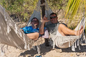 Two men relaxing in side-by-side hammocks under the shade of trees, smiling and enjoying the outdoors. One of them is making a friendly hand gesture while the other lounges comfortably, wearing sunglasses and casual clothing. The setting is a sandy area with flora in the background.