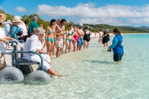 A group of people standing and gathered at the shoreline of a clear, shallow beach. A person in a beach wheelchair dips their feet in the water, while a guide stands in the water, engaging with the group. 